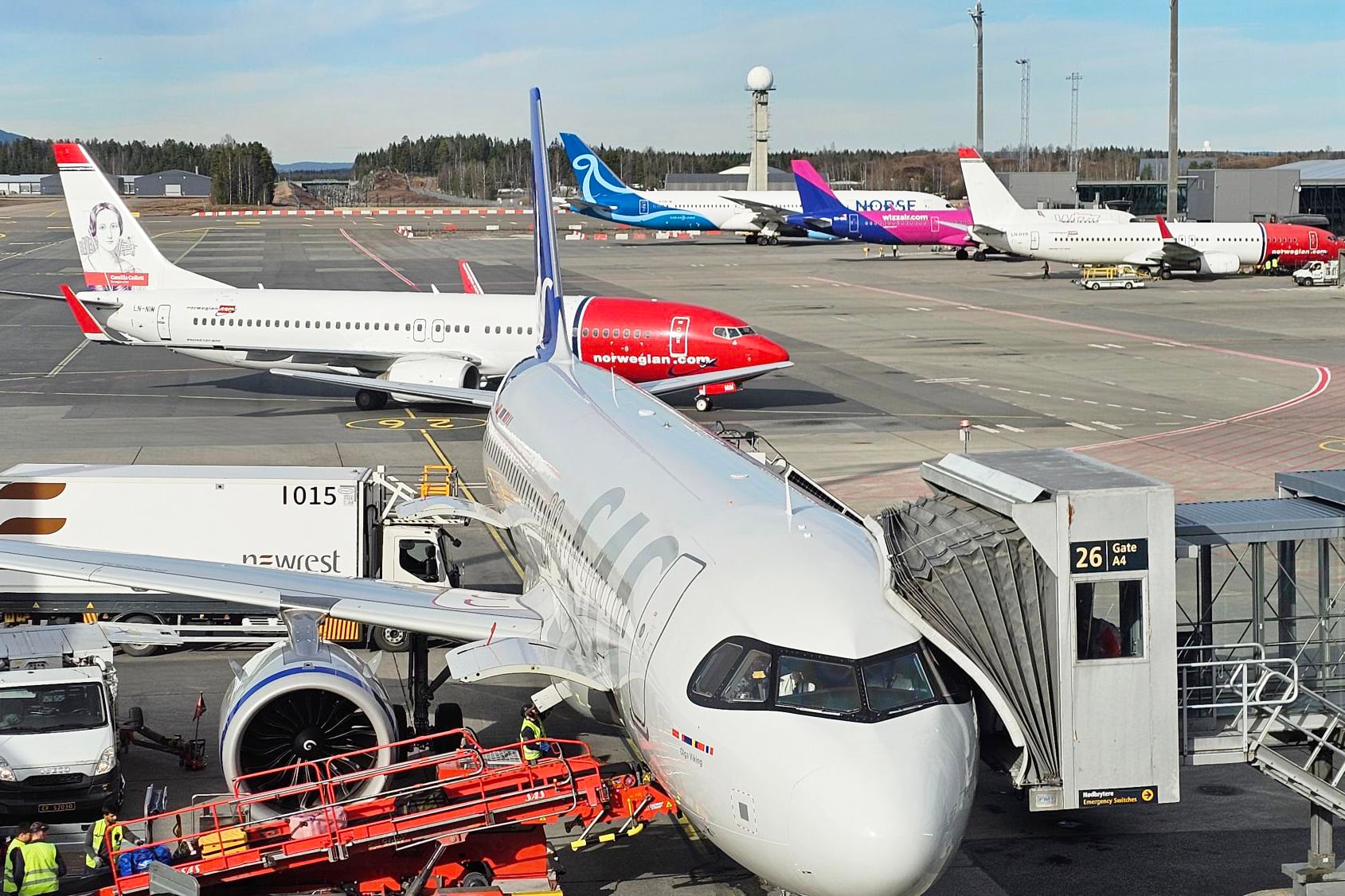 Et passasjerfly fra SAS står parkert ved gate på Oslo lufthavn med passasjerbro tilkoblet. Bakkemannskap og servicekjøretøy arbeider rundt flyet. I bakgrunnen står flere fly fra Norwegian, Norse Atlantic og Wizz Air parkert på flyplassen under klarvær.