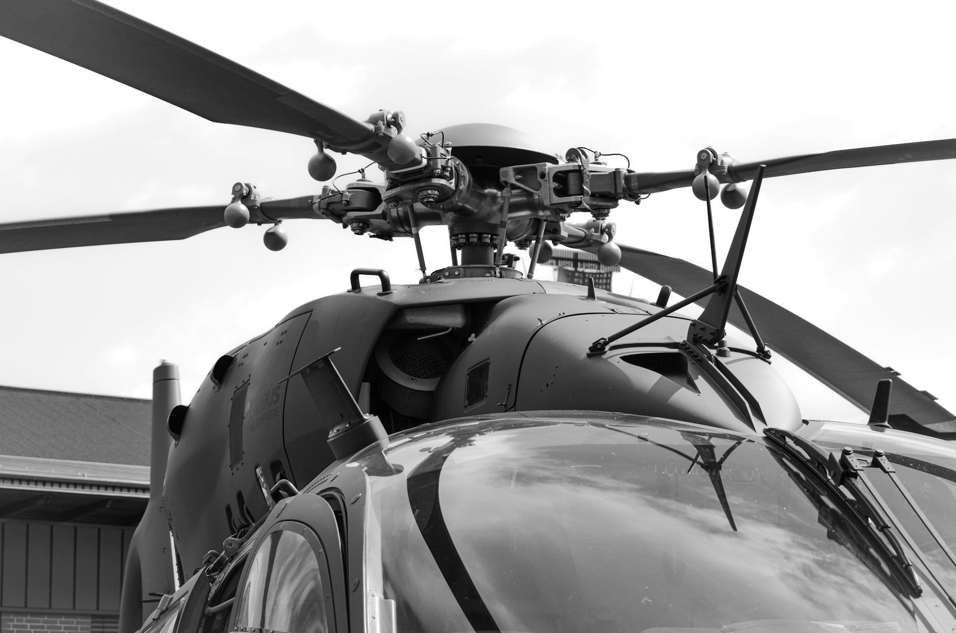 Nærbilde av den øvre delen av et helikopter, med fokus på hovedrotoren, rotorhodet og cockpiten. Bildet er tatt nedenfra og viser de tekniske detaljene i rotorsystemet mot en overskyet himmel. Fotografiet er i svart-hvitt.